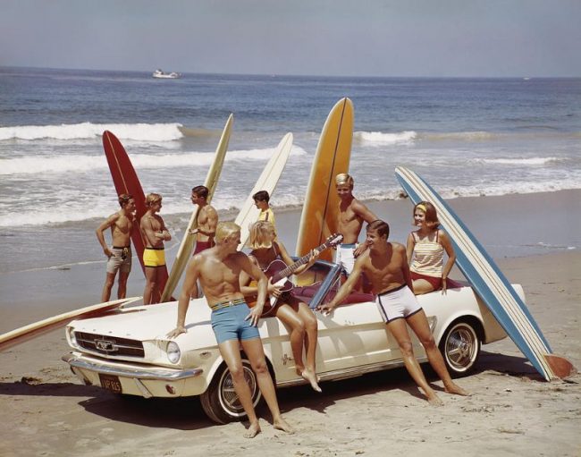 A Group Of Guys And Gals And Their Surfboards Gather Round A Ford Mustang On The Beach, 1964. Photo By Tom Kelley